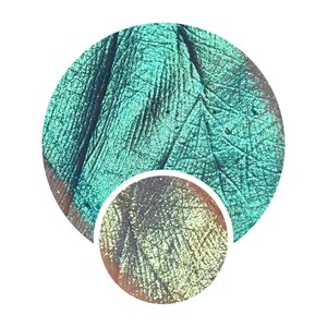 May include: Close-up of a swatch of teal and green iridescent makeup. The makeup has a textured, almost cracked appearance, with a smaller, circular swatch below showing a shift in colour to gold and green.