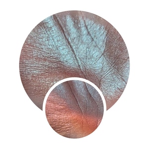 May include: Close-up of a shimmery, iridescent eyeshadow in a light blue and green colour with a copper-toned shimmer. The eyeshadow is shown on a person's skin.
