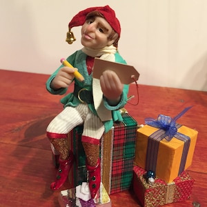 May include: A green and white elf figurine with a red hat and a bell sits on top of a stack of wrapped gifts. The elf is holding a pencil and a small tag. The gifts are wrapped in plaid, orange, and red paper.