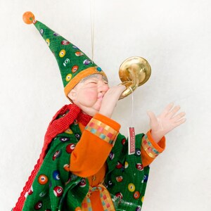 Peut inclure: Une décoration de Noël artisanale et fantaisiste représentant un gnome portant un chapeau vert et orange avec une écharpe rouge et jouant de la trompette dorée. Le gnome porte une chemise à motifs verts et oranges et a une barbe blanche.