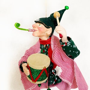 Peut inclure: Une décoration d'elfe de Noël avec un chapeau vert, un manteau rayé rouge et blanc et un tambour vert. L'elfe tient un bâton de tambour et un tuyau vert.