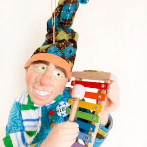 Peut inclure: Une décoration de Noël artisanale représentant un lutin fantaisiste portant un chapeau bleu et marron et une écharpe rayée bleu et blanche. Le lutin tient un xylophone en bois avec un maillet et sourit.
