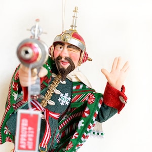 Peut inclure: Une décoration de Noël fantaisiste représentant un homme portant un bonnet de Père Noël rouge et blanc avec une écharpe rayée verte et blanche et tenant un pistolet jouet. La décoration est ornée de rayures de cannes en sucre et d'un flocon de neige avec le numéro 517.