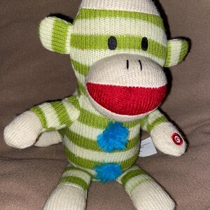 May include: Green and white striped sock monkey plush toy with a red mouth and blue pom-poms. The monkey is sitting on a brown surface.
