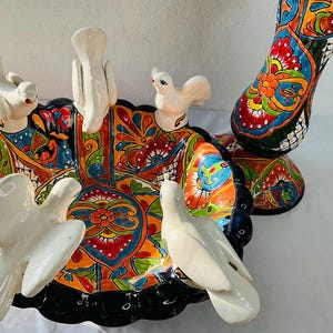 Może przedstawiać: Kolorowa ceramiczna kąpiel dla ptaków z czarną krawędzią i białymi gołębiami. Kąpiel dla ptaków jest ozdobiona wzorami kwiatowymi w odcieniach niebieskiego, czerwonego, żółtego i zielonego.