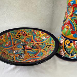 Może przedstawiać: Ręcznie malowana ceramiczna miska do serwowania i wazon dekoracyjny. Owalna miska ma czarną obwódkę, a wazon jest cylindryczny. Oba elementy zdobią skomplikowane wzory kwiatowe i geometryczne w żywych kolorach, takich jak niebieski, zielony, czerwony i żółty.