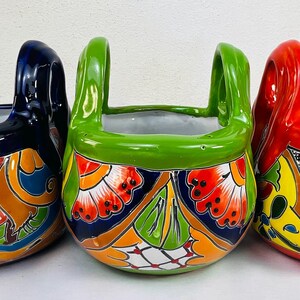 May include: Three brightly coloured ceramic planters with handles. The planters are painted in blue, green, and red, with detailed floral and geometric patterns. The interior of each planter is white.