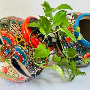 May include: Two large, hand-painted ceramic planters with vibrant floral and geometric patterns in red, blue, orange, and green. One planter has a red handle, the other a blue rim. Green leafy vines cascade from the planters, adding a natural element.