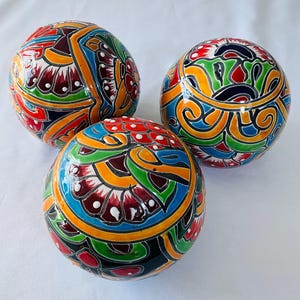 May include: Three spherical objects featuring a glossy finish and intricate hand-painted designs in vibrant shades of red, yellow, blue, green, and black. Each sphere displays complex floral and geometric patterns with raised white dot accents.