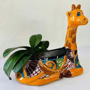 Może przedstawiać: Kolorowa ceramiczna doniczka w kształcie żyrafy z zieloną rośliną w środku. Doniczka jest ozdobiona geometrycznym wzorem w odcieniach pomarańczowego, brązowego, niebieskiego, zielonego i białego.