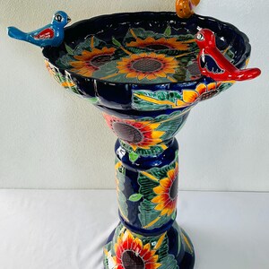 May include: A ceramic birdbath featuring a deep blue glaze decorated with hand-painted yellow and orange sunflowers and green leaves. The basin rim is adorned with three small, colorful ceramic birds in blue, orange, and red.