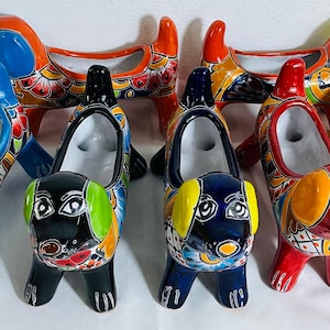 May include: A collection of seven ceramic dog-shaped planters, each featuring vibrant, multi-colored hand-painted floral and geometric patterns. The planters are arranged in two rows, showcasing various colors including blue, black, red, and yellow.