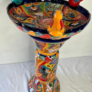 Puede incluir: Un baño de pájaros de cerámica colorido con un pedestal. El baño de pájaros está decorado con un patrón tradicional mexicano Talavera en tonos de azul, amarillo, rojo, verde y blanco. El baño de pájaros presenta tres figuras de pájaros, dos posadas en el borde y una en el lateral.