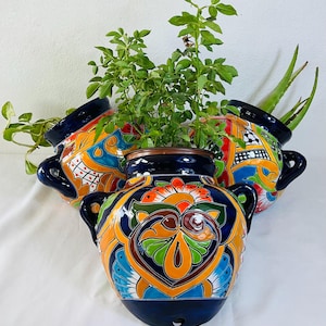 May include: Three ceramic wall planters featuring a glossy dark blue base with vibrant hand-painted patterns in orange, green, yellow, and white. Each rounded vessel includes two side handles and a small drainage hole at the bottom center.