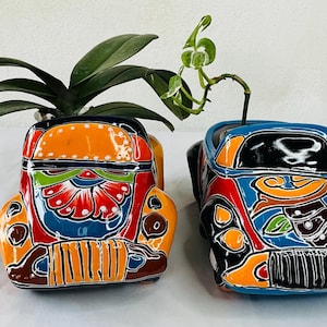 May include: Two colorful ceramic planters shaped like vintage cars. The planter on the left is orange, yellow, red, blue, and green. The planter on the right is blue, black, orange, yellow, and green.