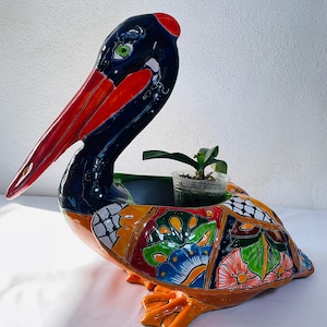 May include: A ceramic pelican planter with a vibrant, hand-painted design. The pelican features a red beak, orange feet, and a colorful body with floral and geometric patterns. A small plant sits inside the planter, adding a touch of greenery.