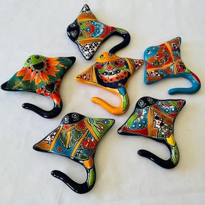 May include: Six ceramic stingray-shaped hooks, each with a unique hand-painted design. The hooks feature vibrant colours including blue, orange, green, and yellow. The curved tail of each hook is designed for hanging items. The background is white.
