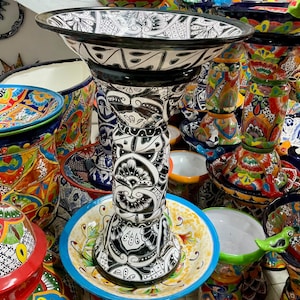 Puede incluir: Un bebedero para pájaros de cerámica blanco y negro con intrincados diseños florales. El bebedero para pájaros está rodeado de otros artículos de cerámica coloridos, incluyendo cuencos y macetas.