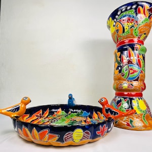 Puede incluir: Un vibrante bebedero para pájaros y un pedestal decorativo de cerámica pintados a mano. El bebedero es azul oscuro con detalles en naranja y amarillo, con tres pequeñas figuras de pájaros. El pedestal es alto, con patrones coloridos y una parte superior acampanada.