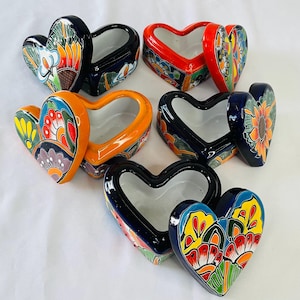 Puede incluir: Cinco cajas de cerámica en forma de corazón con tapas. Las cajas están decoradas con motivos florales en tonos azules, rojos, amarillos, naranjas y verdes. Las tapas también están decoradas con motivos florales.