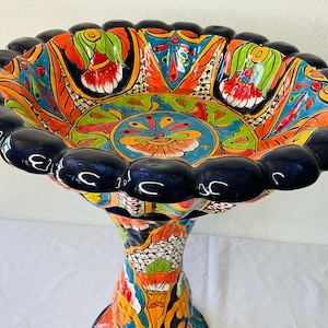 Puede incluir: Un baño de pájaros de cerámica colorido con un borde festoneado y un diseño floral detallado. La base es una columna alta y retorcida con un patrón floral similar.