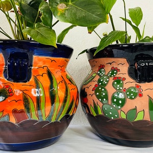 Könnte beinhalten: Zwei Keramik-Pflanztöpfe mit handgemalten Wüstenmotiven. Ein Topf zeigt orange und blaue Farbtöne mit Kakteen und grünen Pflanzen. Der andere Topf hat einen beigefarbenen Hintergrund mit grünen Kakteen und Pflanzen.