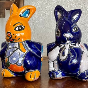 May include: Two ceramic rabbit figurines, one yellow and blue, the other blue and white, with painted details. Both have a blue and white patterned design on their bodies.