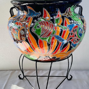 May include: A large, round, dark blue ceramic planter with two handles, featuring a vibrant, hand-painted underwater scene with colorful fish, orange sunburst patterns, and green accents, resting on a black metal tripod stand with curved legs.