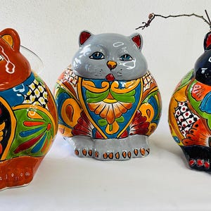 May include: Three ceramic cat figurines with vibrant, hand-painted designs. The cats are in shades of orange, gray, and black, with intricate patterns in blue, green, and orange. Each cat has blue eyes and a glossy finish, adding a decorative touch.