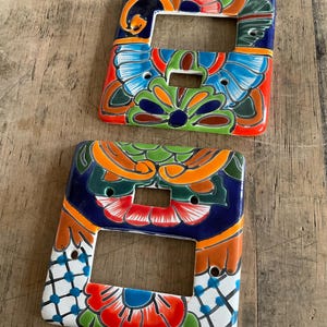 Könnte beinhalten: Zwei farbenfrohe Keramik-Lichtschalterabdeckungen. Jede Abdeckung hat eine rechteckige Form mit einer zentralen Öffnung und ist mit einem lebendigen Blumendesign in Rot-, Blau-, Grün- und Orangetönen verziert. Sie liegen auf einer Holzoberfläche.