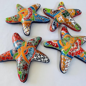 May include: Four colorful, ceramic starfish decorations. Each starfish features a unique hand-painted design with vibrant hues of red, orange, blue, green, and white. The starfish have a glossy finish and are arranged on a white surface.