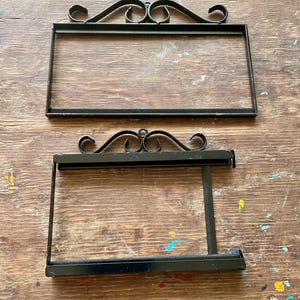 Puede incluir: Dos soportes de letreros de metal negro con adornos decorativos en la parte superior. Los marcos rectangulares están vacíos y uno tiene un brazo de soporte. Los soportes de letreros se muestran sobre una superficie de madera envejecida.