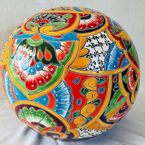 May include: A large, spherical ceramic orb featuring a vibrant, hand-painted Talavera-style pattern. The surface is decorated with intricate floral motifs, geometric shapes, and a bold color palette of red, yellow, blue, green, and black.