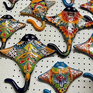 May include: A collection of hand-painted ceramic stingray wall hooks. Each hook is decorated with a unique, colourful design, featuring shades of blue, orange, green, and yellow. The hooks have a curved tail and are displayed on a white surface with small black dots.