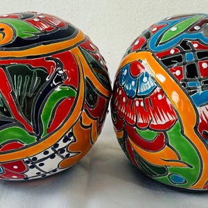 May include: Two decorative ceramic spheres with intricate, colorful floral designs. The spheres feature a vibrant palette of red, orange, green, blue, and white, with black outlines. The handcrafted spheres are likely for home decor.