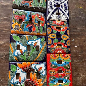 Puede incluir: Una colección de coloridas cubiertas de cerámica para interruptores de luz. Cada cubierta presenta un diseño único pintado a mano con colores vibrantes, incluyendo patrones florales, de peces y abstractos. Las cubiertas son rectangulares con aberturas para interruptores.