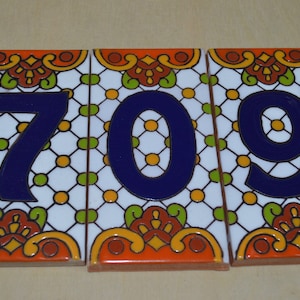 Op de afbeelding: Drie decoratieve keramische tegels met het nummer 709. De tegels hebben een witte achtergrond met een rasterpatroon, blauwe cijfers en oranje, gele en groene decoratieve randen. Elke tegel is ongeveer 15 cm breed.