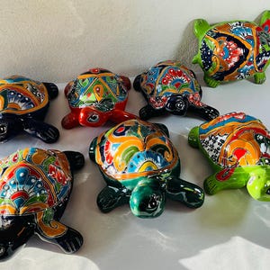 May include: A collection of ceramic turtle figurines. Each turtle has a brightly coloured shell with intricate patterns in shades of blue, red, orange, green, and yellow. The turtles' bodies are various colours, including black and green.