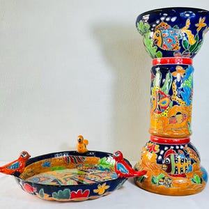 Może przedstawiać: Ręcznie malowany zestaw ceramiczny składający się z płytkiej misy z trzema małymi figurkami ptaków na krawędzi oraz wysokiego cylindrycznego stojaka. Oba elementy są ozdobione żywymi, wielokolorowymi motywami ryb i roślin wodnych.