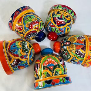 May include: Five ceramic bell-shaped ornaments featuring intricate hand-painted floral and geometric patterns in vibrant shades of red, blue, yellow, orange, and green. Each bell has a solid-colored handle in blue, red, or orange.