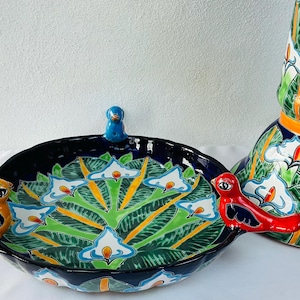 Może przedstawiać: Dekoracyjna ceramiczna patera do serwowania z pokrywką i pasujący wazon dekoracyjny. Patera jest granatowa z kolorowym kwiatowym wzorem i uchwytami w kształcie ptaków. Wazon ma podobny wzór z zielonymi, białymi i pomarańczowymi akcentami.