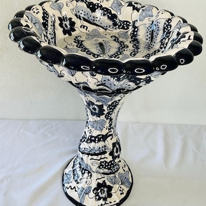 Puede incluir: Un bebedero para pájaros de cerámica blanca con un borde negro y un diseño floral azul y blanco. El bebedero tiene una parte superior ancha y festoneada y una base alta y delgada.