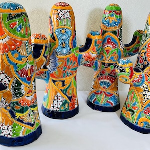 May include: Four ceramic cactus sculptures featuring vibrant, hand-painted floral and geometric patterns in shades of orange, blue, green, yellow, and red. Each piece has a glossy finish, a wide base, and two side arms extending outward.