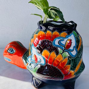Può includere: Un vaso in ceramica a forma di tartaruga con una base blu scuro lucida e un vivace design dipinto a mano con girasoli arancioni, foglie verdi e dettagli floreali bianchi. La tartaruga presenta una testa di colore arancione brillante.