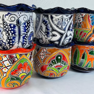 May include: A collection of five ceramic planters with scalloped rims, featuring hand-painted designs in vibrant colours including orange, green, blue, and white. The patterns include butterflies, floral motifs, and cactus illustrations.