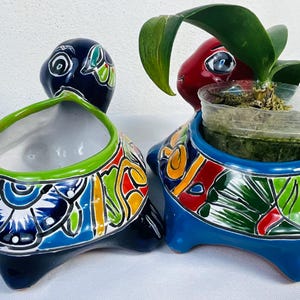 Może przedstawiać: Dwie ceramiczne donice w kształcie żółwi. Jedna jest granatowa z zieloną obwódką i kolorowymi kwiatowymi wzorami. Druga jest niebieska z czerwoną głową i zawiera małą roślinę. Obie mają szczegółowe, ręcznie malowane wzory.