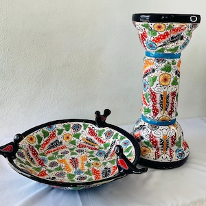 Puede incluir: Un cuenco y un pedestal decorativos de cerámica con un diseño de mariposas y flores. El cuenco ovalado tiene un borde negro y asas con forma de pájaro. El pedestal, alto y cilíndrico, tiene una parte superior e inferior ensanchadas. Ambas piezas presentan un patrón colorido.