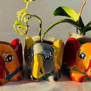 May include: Three colorful elephant-shaped planters. The planters are painted in shades of orange, yellow, red, and blue. Each planter has large ears and a long trunk. The planters are holding small plants.