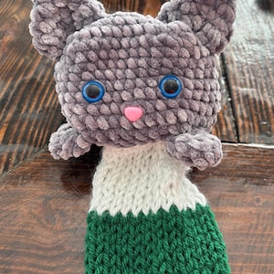 May include: A gray crocheted plush toy with blue eyes and a pink nose. The toy is wearing a white and green striped knitted hat.