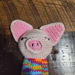 May include: A crocheted pig toy with pink ears and a pink snout. The pig is wearing a colorful striped scarf.